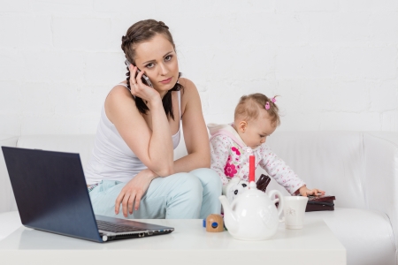 Small child and busy young woman with notebook and phone sit on the sofa in the room.の写真素材