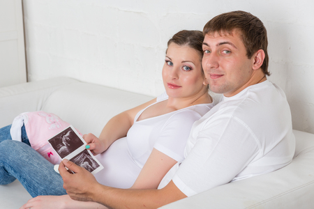 Happy pregnant family with ultrasound picture of their future baby sitting on a sofa of the house.の写真素材