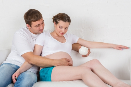 Young pregnant woman and her husband with cosmetic cream sit on a sofa in the room  Concept of body care の写真素材