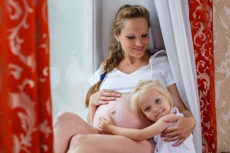 Pregnant woman with her little daughter  sit near window in the room. Happy family.
