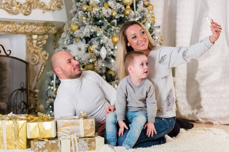 Happy family with a mobile phone and gift boxes sit near a Christmas tree in the house.  Merry Christmas and Happy New Year.の写真素材