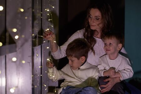 Happy family are sitting near window decorated Christmas garland at home. Christmas and New Year holiday. Beautiful young mother with two sons.の写真素材