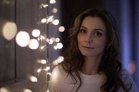 Happy young beautiful woman is standing near window decorated Christmas garland at home. Christmas...