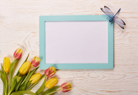 Blank sheet in green frame, bouquet of tulips and dragonfly on a wooden background. Top view with...
