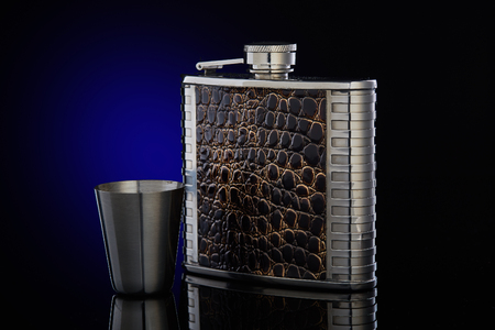 Hip flask from stainless steel with brown leather  for whisky or cognac and steel drinking glass on a dark blue background with copy space.の写真素材