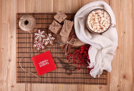 Glass mug with hot cocoa and marshmallow, gift boxes and greeting card on wooden background. ...