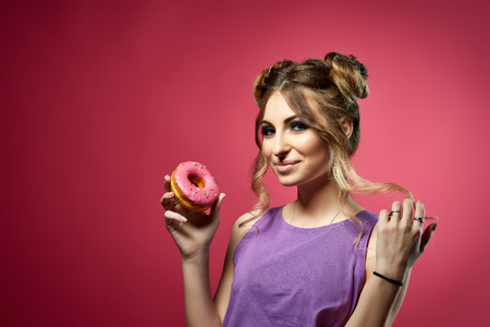 Cheerful young woman in bikini and top stands with tasty donut on a pink background. Pin up style.の写真素材