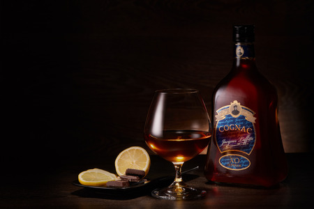 Bottle of cognac Jacques Laffitte XO Superior, glass with cognac and plate with pieces of lemon and chocolate stands on a table on a dark wooden background.のeditorial素材
