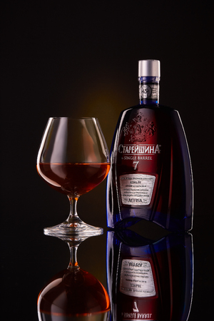 Bottle of cognac Starejshina and glass with cognac on a black background with reflection.のeditorial素材