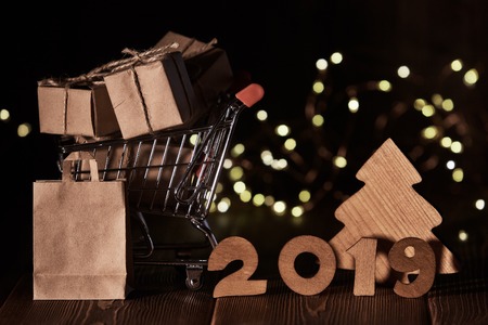 Wooden fir tree, gift boxes, shopping trolley and text 2019  from wooden figure on dark wooden background with LED light garland. Horizontal view. New Year and Christmas.の写真素材