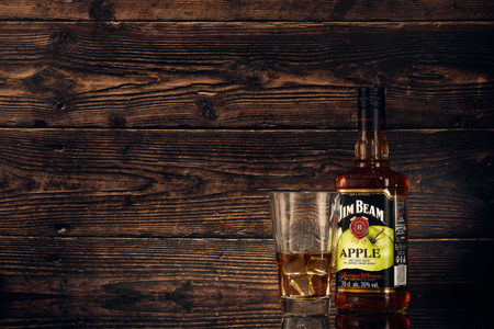 Still life with bottle of Jim Beam Apple (apple liqueur infused with kentucky straight bourbon), glass with bourbon, ice  on a brown wooden background with copy space.のeditorial素材