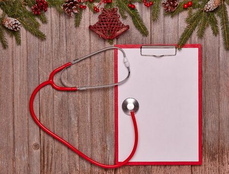 Red stethoscope, blank sheet on clipboard, fir tree  branches and Christmas ornaments on wooden background. Top view with copy space. Medical concept. Greeting card. New Year and Christmasの写真素材