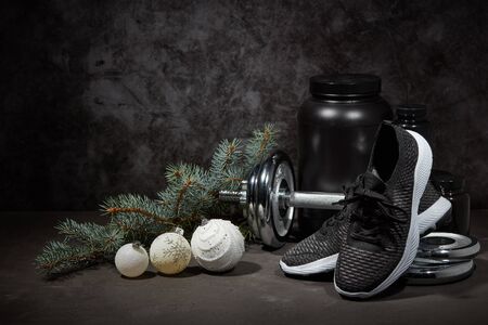 Sports nutrition (supplements), dumbbell, sneakers, branch of fir and Christmas ornaments on dark grey background with copy space. Fitness, sport or healthy lifestyle concept. New Year and Christmas.の写真素材