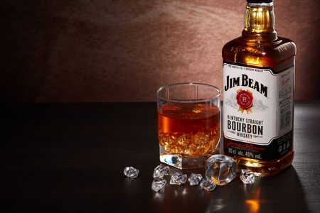 St.Petersburg, Russia - December 2019 - Bottle of Jim Beam bourbon whiskey and glass with drink and ice  on wooden table on brown background with copy space. Kentucky straight bourbon whiskeyのeditorial素材