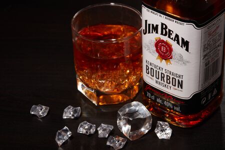 St.Petersburg, Russia - December 2019 - Bottle of Jim Beam bourbon whiskey and glass with drink and ice  on wooden table on brown background. Kentucky straight bourbon whiskeyのeditorial素材