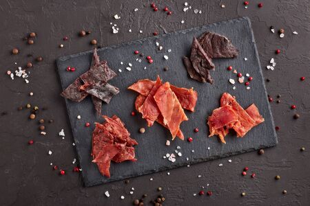 Jerky. Set of various kind of dried spiced meat on black stone cutting board on dark gray background. Top view.  Snack for beer.の写真素材