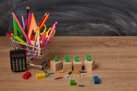 Shopping trolley with stationery accessories and calculator on wooden table on background of school...