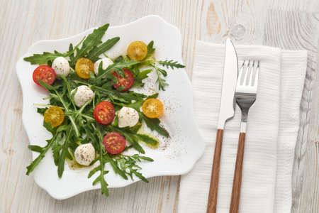 White plate with dietary salad from mozzarella cheese, cherry tomatoes and leaves of arugula on light wooden background , table setting.の写真素材