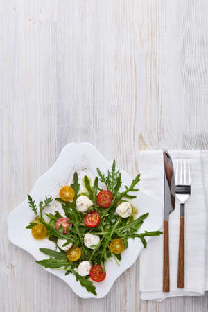 White plate with dietary salad from mozzarella cheese, cherry tomatoes and leaves of arugula on light wooden background , table setting. Top view with copy space.の写真素材