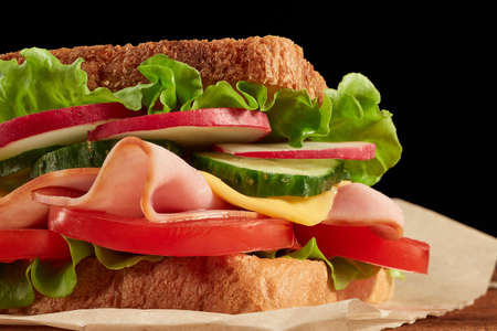 Appetizing sandwich from ham, cheese and vegetables serving on parchment paper is laying on wooden...