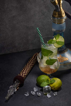 Hookah (shisha) and glass of Mojito cocktail with lime and ice on dark grey background. Weekend or...