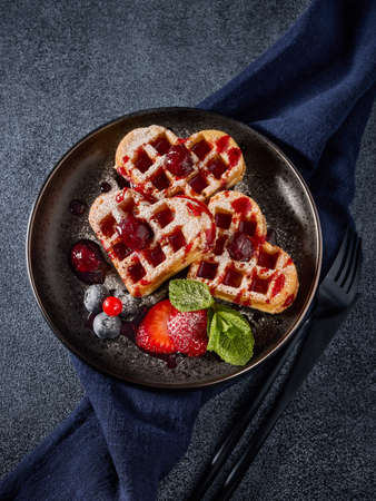 Delicious waffles heart shape with  powdered sugar and berries on ceramic black plate on grey...
