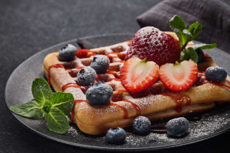 Delicious waffles with  powdered sugar and berries on ceramic black plate on grey background. Sweet...