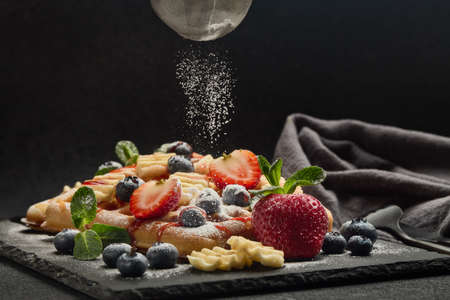 Delicious waffles with berries, mint, powdered sugar, sweet sauce on slate serving board on dark background.  Dessert. Serving food. Process of sprinkling icing sugar over wafflesの写真素材