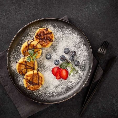 Russian cottage cheese pancakes (Syrniki) with berries, powdered sugar, chocolate sauce on ceramic  plate on black background. Top view. Dessert. Serving foodの写真素材