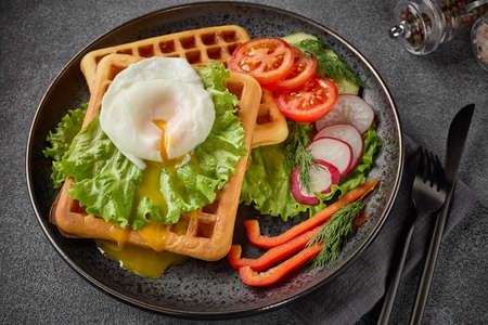 Delicious waffles with poached egg and fresh vegetables on ceramic black plate on slate grey background. Top view. Serving food. Table settingの写真素材