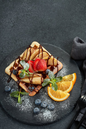 Delicious waffles with caramelized pear, berries, mint, powdered sugar and sweet sauce on black serving board on grey background. Dessert. Serving food. Table settingの写真素材