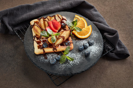 Delicious waffles with caramelized pear, berries, mint, powdered sugar and sweet sauce on black serving board on dark brown background. Dessert. Serving food. Top viewの写真素材