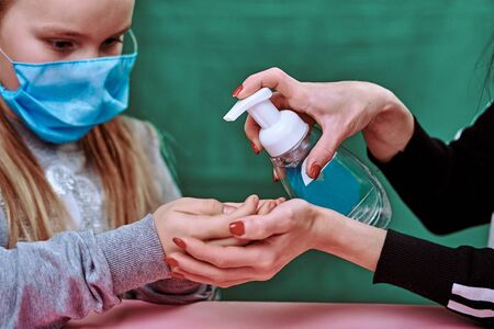 mom disinfects her baby 's hands. the child is wearing a blue medical mask. quarantine. disinfectant fights bacteria. green background and pink table. covid-19. protect the healthの写真素材