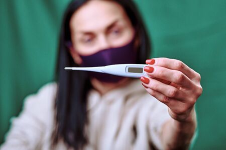 a brunette girl wearing a purple medical mask shows a white electronic thermometer into the camera....