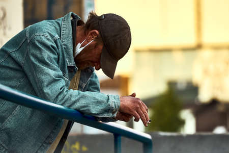Kiev, Ukraine, 04.08.2020: A man wearing a medical mask poor stands and smokes lowered head. It became even more difficult to live in quarantine. street photo. covid-19のeditorial素材