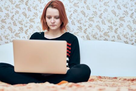 A girl in a striped jacket holds a laptop in her hands and sits at home in quarantine and prints in a computer. Entertained, worked and studied at home. covid-19. stay at home. work home. without make-upの写真素材