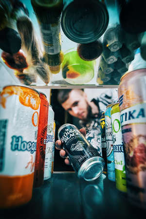Kiev, Ukraine, 20.05.2020: The guy gets a can of beer from the fridge, where other types of beer are located. The top row of the refrigerator is clogged with food. advertizing photo.のeditorial素材