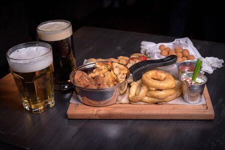 Two bakals of beer, dark and light, snacks for beer. Fried chicken legs, onion rings, cheese balls, fried vareniki and sauces on a tree board. for the menu of cafe. wooden table. side view.の写真素材
