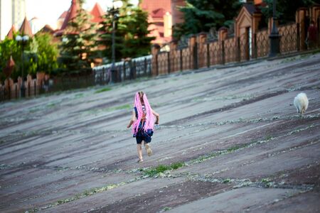 A girl with pink dreads runs down the street with a small white dog. rear view. concrete slabs, street.の写真素材
