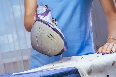The girl holds an iron on an ironing board with a ferry and irons things. Cleaning service of the apartment. Blue T-shirt and young model who smiles. macro. to raise the equipmentの写真素材