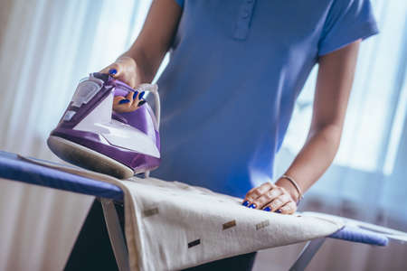 The girl holds an iron on an ironing board with a ferry and irons things. Cleaning service of the apartment. Blue T-shirt and young model who smiles. macro. to raise the equipmentの写真素材