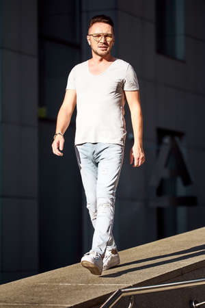a stylish guy with glasses walks along the sidewalk along an office building. strong male gaze. light jeans and a fashionable gray T-shirt. sunlightの写真素材