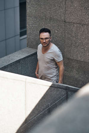 a stylish guy with glasses looks at the camera, goes down the steps to the office. strong strict male gaze. fashionable gray T-shirt. sunlight. photo art.の写真素材