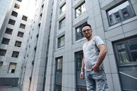 a stylish guy with glasses looks into the camera against the background of an office building in gray, hands in pockets. strong strict male gaze. a fashionable gray T-shirt and light jeans. portraitの写真素材