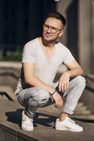 a stylish guy with glasses sat on the sidewalk, smiling a little and looking away. strong male gaze. light jeans and a fashionable gray T-shirt. sunlightの写真素材