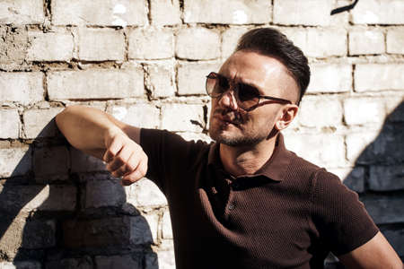 a stylish guy with glasses wrapped himself on a stone wall with an elbow and looks away. strong and strict male gaze. brown T-shirt with collar and fashionable beige trousers. sunlight. half-length portraitの写真素材
