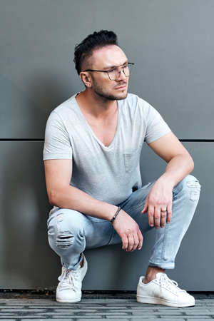 a stylish guy with glasses sat down near a gray wall and looks away. strong and strict male gaze. light jeans and a fashionable gray T-shirt. sunlight. half-length portraitの写真素材