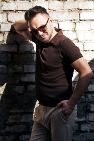 stylish guy with glasses stands near a stone wall and looks away. thought. strong and strict male gaze. brown T-shirt with collar and fashionable beige trousers. sunlight.の写真素材