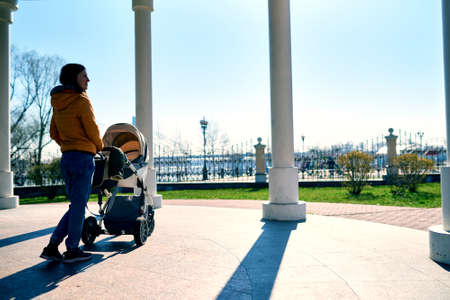 A young mother walks with a stroller and a child on the street to the park and drinks coffee. Leisure in nature. Sunny weather. Architecture and nature. Orange jacket. Healthy lifestyleの写真素材