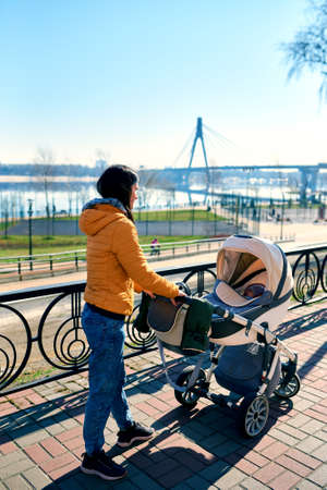 A happy young mother rolls a stroller with a child on the street to the park. leisure in nature. Sunny weather and beautiful scenery. Orange jacket. Healthy lifestyleの写真素材